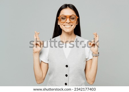 Young latin woman she wears white t-shirt gray vest glasses waiting for special moment, keeping fingers crossed, making wish, eyes closed isolated on plain grey background studio. Lifestyle concept