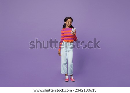 Full body happy little kid teen girl 15-16 years old wear striped orange sweatshirt use mobile cell phone chatting online with friends isolated on plain purple background. Childhood lifestyle concept