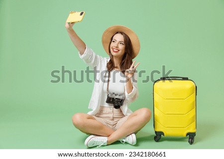 Full length traveler tourist woman in casual clothes hat hold suitcase do selfie mobile phone show v-sign sit isolated on green background Passenger travel abroad weekends Air flight journey concept
