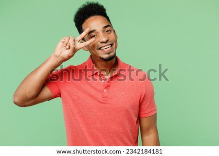 Young smiling man of African American ethnicity wears pink t-shirt showing cover eye with victory sign look camera isolated on plain pastel light green background studio portrait. Lifestyle concept