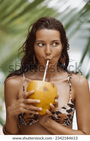 Beach fun. Funny portrait of a young adult bikini beach woman in a leopard print swimsuit with fresh coconut near the sea under a palm tree