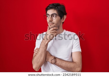 Young hispanic man standing over red background looking confident at the camera smiling with crossed arms and hand raised on chin. thinking positive.  Royalty-Free Stock Photo #2342189511