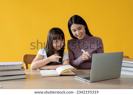 An adorable young Asian girl enjoys learning English online on a laptop with a female teacher, sitting at a table together. isolated yellow background. Royalty-Free Stock Photo #2342195319