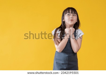 A happy and adorable little Asian girl in a cute jeans dress is standing against an isolated yellow background in a cute pose. surprised, saying wow, excited, amazed, can't wait for the good news