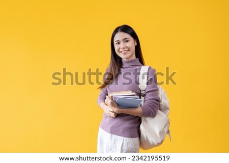An attractive young Asian female college student with a backpack holding her school books, smiling and looking at the camera while standing against an isolated yellow background.
