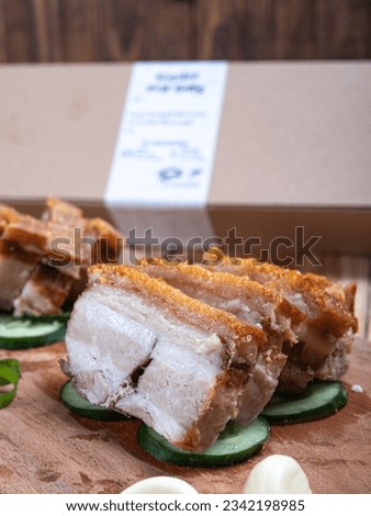 The roasted pork is neatly arranged in a row on a wooden cutting board, garnished with cucumbers, and surrounded by various spices, isolated with a grayish wooden table background.