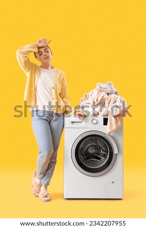 Tired young woman standing near washing machine with pile of dirty clothes on yellow background