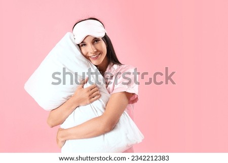 Young woman with sleeping mask hugging pillow on pink background