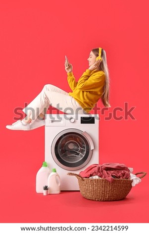 Pretty young woman sitting on washing machine and listening to music against red background