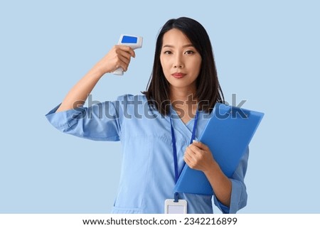 Female Asian medical intern with infrared thermometer and clipboard on blue background