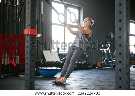 Full-length image of mature woman training in gym, doing exercises with gymnastics rings. Hand pull-ups exercises. Strength. Concept of active, sportive and healthy lifestyle, fitness, body care, ad
