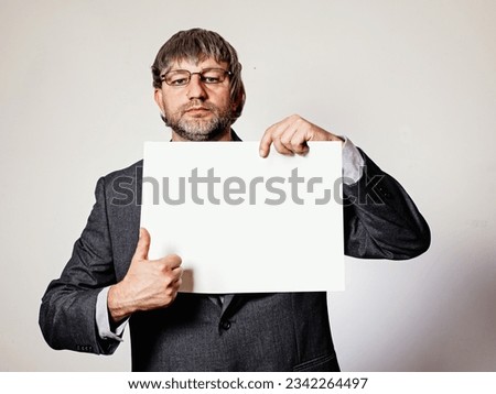Man holding white blank message board. Grey wall background. The model is in his 40s with grey beard and hair wearing business suit and glasses. Manager message to his team or advertisement concept.