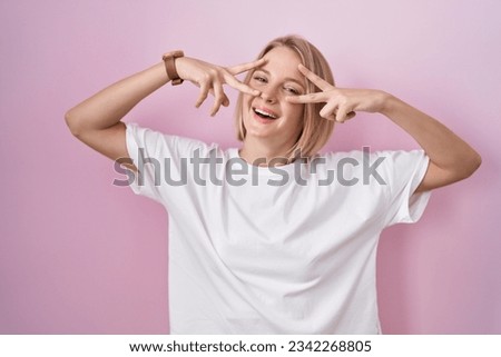 Young caucasian woman standing over pink background doing peace symbol with fingers over face, smiling cheerful showing victory 
