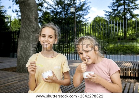 portrait of two cute blonde girls, they eat cold ice cream from paper cups. A walk in the park, summer holidays and fun