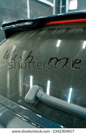 A close-up of a wash me sign on the dirty rear window of a luxury blue car in front of car wash