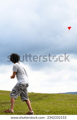 Male with curly hair wearing shorts is flying a red kite on green meadow. Turkish man flying a kite. People, human, model. Vertical photo. Childhood idea concept. Enjoying freetime.  Royalty-Free Stock Photo #2342332153