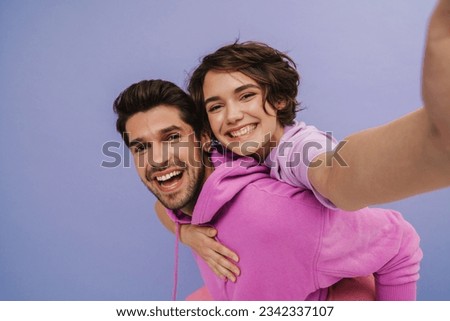 Cheerful brown-haired man carrying his smiling girlfriend on his back while she taking selfie isolated over purple studio background