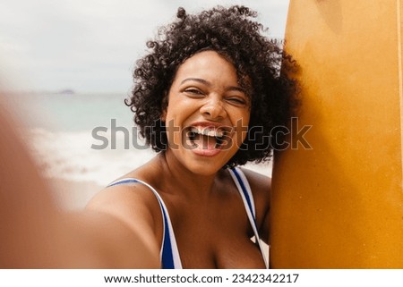 Happy young woman with curly hair taking a selfie with her surfboard on the beach. Excited female surfer capturing the moments of her summer vacation before going on an adventurous surfing trip.