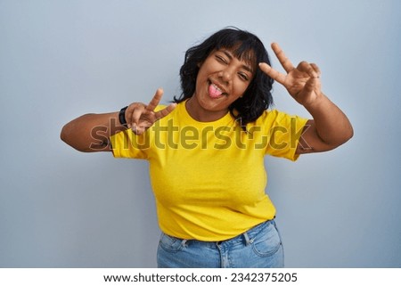 Hispanic woman standing over blue background smiling with tongue out showing fingers of both hands doing victory sign. number two. 