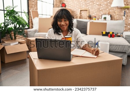 Young beautiful latin woman using laptop writing on notebook at new home