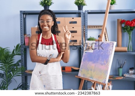 Young african american with braids at art studio painting on canvas smiling with happy face winking at the camera doing victory sign. number two. 