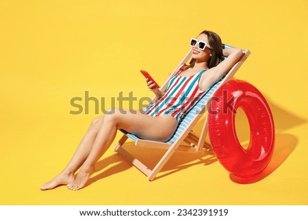 Full body length fun happy young woman wear red blue swimsuit sit on wooden chair hold use mobile cell phone isolated on vivid yellow color background studio Summer hotel pool sea rest sun tan concept