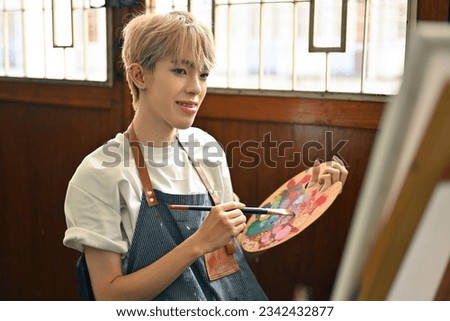 Portrait image of an Asian LGBT teenage Artist student with colored hair doing his artwork, painting color on a canvas in the studio workshop.