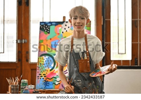 Portrait image of a Young Asian gay Artist boy with colored hair posing with his artwork and looking at the camera inside the studio workshop.