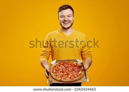 Excited man in yellow sweater holding cardboard flat box with pizza isolated on orange background. Food delivery concept