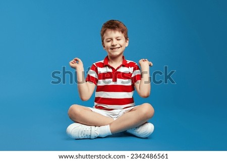 Peaceful cute little boy holding fingers in mudra gesture and meditating with closed eyes while sitting in lotus pose, feeling calm positive and relaxed, yoga practice, isolated on blue background