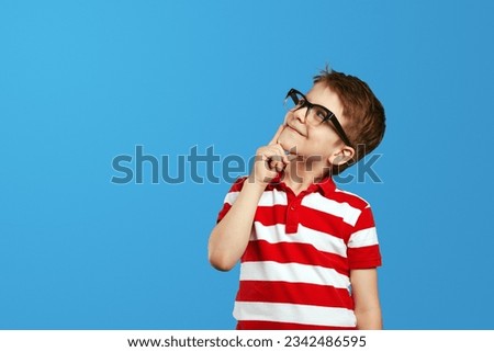 Smart wunderkind in nerdy glasses and red striped polo shirt touching cheek and looking up while thinking against blue background.