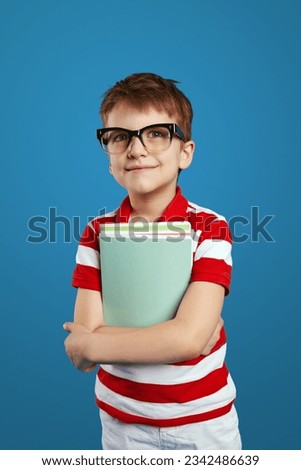 Clever schoolboy in nerdy eyeglasses smiling and holding textbooks while looking away, standing isolated over blue background. Back to school concept.