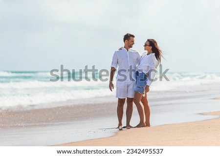 Happy couple in love on the beach vacation walking together at sunset. Young woman in white dress and caucasian man in white shorts. Royalty-Free Stock Photo #2342495537