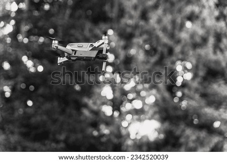 A grayscale of a drone in the air, sunlit trees blurred background