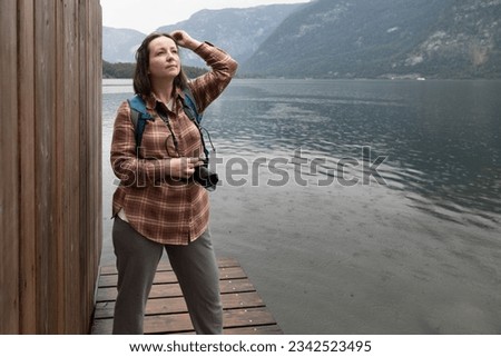 Young female photographer standing on the shore of lake with camera in her hands and looking into the distance, Austria