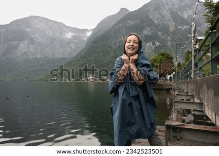 Young woman in a raincoat standing on the shore of a lake in the rain and smiling at the camera, Hallstatt, Austria
