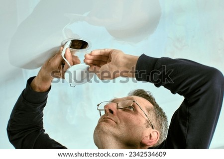 Installing spotlight in stretch ceiling, an elderly white electrician inserts an LED lighting fixture into hole. Royalty-Free Stock Photo #2342534609