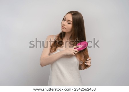Portrait of positive girl in nightgown brushing her beautiful shiny hair with hairbrush. Attractive woman isolated on gray background. Hair care concept