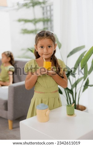 Children's Look: Young Girl with Wooden Cup, Engaging with Montessori Playset in Green Dress on a White Background