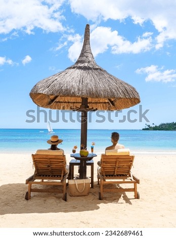 Man and Woman on a tropical beach with Chairs And Umbrella In Tropical Beach. Beautiful tranquil white sand beach with two chairs and thatched umbrella, couple drinking cocktails on the beach by ocean