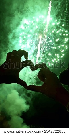 a photography of a person making a heart with their hands, fireworks in the sky with two hands making a heart shape.