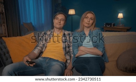 The couple is sitting on the sofa and watching TV. A man changes channels using a remote control. The woman sits with a bored expression on his face. Nothing interesting.