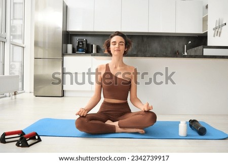 Smiling girl athlete, sportswoman doing yoga at home in activewear, sitting on yoga rubber mat in lotus pose, meditating, practice mindfulness exercises.