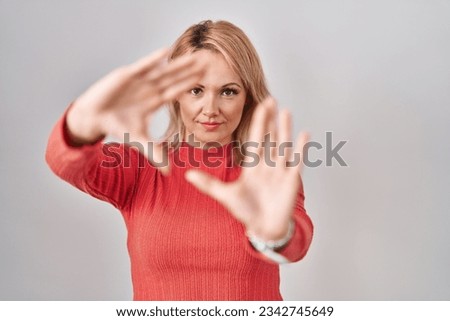 Blonde woman standing over isolated background doing frame using hands palms and fingers, camera perspective 