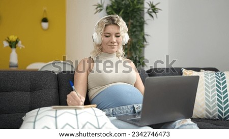 Young pregnant woman using laptop and headphones writing on notebook studying at home