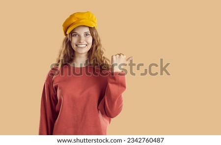 Fashion Model in pastel colors and trendy beret isolated on light brown background. Woman Smiling broadly with teeth and pointing her finger at blank space for promotion and product placement.