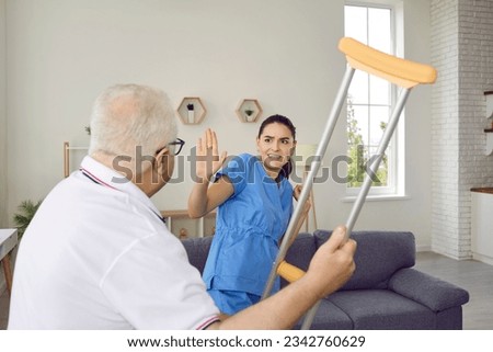 Retired old senior man patient gets very angry, aggressive and dangerous, shows bad attitude toward workers, behaves like crazy psycho, threatens to hit young scared intern volunteer nurse with crutch Royalty-Free Stock Photo #2342760629