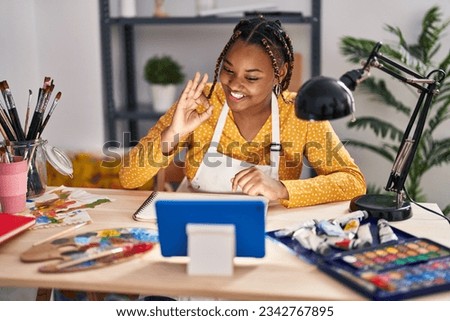 African american woman with braids sitting at art studio painting looking at tablet doing ok sign with fingers, smiling friendly gesturing excellent symbol 