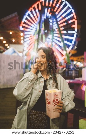 A young lady in a good mood with a popcorn cup in the night at luna park in Santa Monica in front of unfocused illuminations of a Ferris wheel. Santa Monica. California. Royalty-Free Stock Photo #2342770251