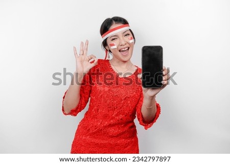A cheerful Asian woman wearing red kebaya and headband, showing her phone while gesturing OK sign with her fingers, isolated by white background. Indonesia's independence day concept